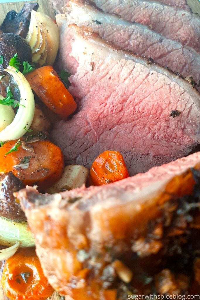 Garlic and Herb Bottom Round Roast Beef on a cutting board, several medium rare slices displayed. Surrounded by roasted carrots, parsnips, onions, and mushrooms. Sprinkled with fresh parsley. Pinterest image. Sugar with Spice Blog.